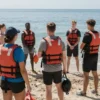 A group of diverse people wearing life jackets are standing on a sandy beach near calm water. Some are holding rescue equipment. The scene is sunny and bright. Focus on safety and preparedness for water activities.