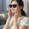 A woman applying sunscreen to her face outdoors on a sunny day. She is wearing a light-colored t-shirt and sunglasses. The background shows a blurred cityscape.
