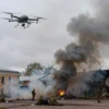 A dramatic scene of a drone attack on the historic city of Kronstadt, with smoke rising from damaged buildings, military personnel taking cover, and a small unmanned aerial vehicle visible in the sky against a cloudy overcast sky, no text, no numbers, no logos
