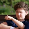A young boy, around 7 years old, sitting on a park bench, gently holding his arm. His arm has a bandage on it. He has a slightly concerned but calm expression. Soft, natural lighting. Background shows a blurred park scene with trees and greenery.
