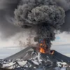 A powerful volcanic eruption at Shiveluch volcano in Kamchatka, with a massive plume of ash rising up to 12 kilometers into the sky, dark clouds spreading across the atmosphere, snow-capped mountain terrain below, and distant smoke-filled skies under a gray daylight