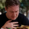 A person feeling overheated while eating a large amount of food outdoors on a hot summer day. They are clutching their stomach, sweating, and looking distressed. Background shows a sunny park with blurred trees and people enjoying the summer. Focus on the person's discomfort.
