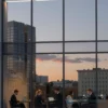 A modern office building in Moscow, Russia, with large glass windows reflecting the city skyline at sunset. Several people in business attire are working on laptops inside the building. Focus on the architectural details and the vibrant colors of the sunset.