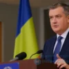A photorealistic image depicting a serious-looking man, Vol, speaking at a press conference. He is wearing a dark suit and tie, and standing behind a podium with a Ukrainian flag partially visible in the background. The setting is a modern press room.