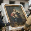 A painting by Roubens, slightly damaged, being carefully transported by rescue workers. The painting is wrapped in protective material. Rescue workers are wearing protective gear. The background shows a damaged building, indicative of war damage. Focus on the painting and the rescue operation.