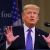 Donald Trump standing confidently at a podium, gesturing with his hands, with a blurred background suggesting a press conference or official event. Focus on his upper body and facial expression.