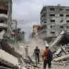 A wide shot of a devastated city in Venezuela after an earthquake. Buildings are collapsed, debris is scattered everywhere, and dust fills the air. Rescue workers in hard hats and protective gear are seen searching through the rubble. The sky is overcast and grey.
