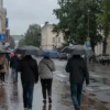 A street scene in Izhevsk on June 14th, showing a rainy day with people walking under umbrellas. Buildings and trees are visible in the background. The overall mood should be slightly melancholic but not depressing.