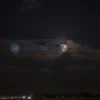 A photo-realistic image depicting the night sky over Voronezh, Russia, during a black moon. The moon should be a dark, almost black orb, partially obscured by clouds. City lights should be visible in the distance, creating a contrast with the dark sky. Stars should be visible.