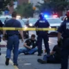 A chaotic urban scene in the United States with multiple people on the ground, emergency responders arriving, and police tape cordoning off an area, all under dim daylight with distant sirens visible, no weapons or blood shown