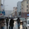 A wide shot of a city street in Chelyabinsk Oblast, Russia, during a rainy morning. People are walking with umbrellas, cars are driving with headlights on, and the buildings have a slightly wet, reflective appearance. Focus on the urban environment and the atmosphere of a rainy workday.