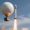 A high-altitude balloon launching a rocket into the stratosphere. The balloon is large and white, with scientific instruments attached. The rocket is sleek and metallic, ascending rapidly against a backdrop of blue sky and wispy clouds. The Earth's curvature is subtly visible on the horizon. Focus on the launch sequence and the contrast between the balloon and the rocket.