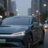 A modern, sleek car parked on a city street at dusk. The car is a dark blue sedan with sharp lines and LED headlights. The background shows blurred city buildings and streetlights.