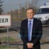 A Russian official in a formal suit standing beside a pothole-filled road near a sign reading 'Пер' (Per), with a concerned expression and a government vehicle parked nearby