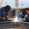 A construction site in Chelyabinsk Oblast, Russia, with several welders working on a large metal structure. Focus on the welders' protective gear and the sparks flying from their welding torches. Include construction equipment like cranes and scaffolding in the background. The scene should convey a sense of industry and economic growth.