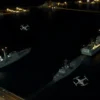 A nighttime aerial view of US Navy ships at a naval base in the Persian Gulf. Several small drones are depicted in flight towards the ships, with trails of light indicating their movement. The scene should convey a sense of tension and military activity. Focus on the ships and drones, with the surrounding environment blurred to emphasize the action.