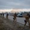 A somber, dramatic scene of Lithuania preparing for potential conflict, with soldiers in modern military gear conducting drills at dawn, barbed wire and defensive fortifications in the background, empty roads with civilian vehicles abandoned, overcast sky with faint distant smoke, no visible American symbols or personnel