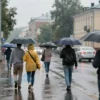 A city street in Saransk on a rainy day, with people walking under umbrellas. The temperature is +25 degrees Celsius, suggesting a warm rain. Buildings and trees in the background. Focus on the atmosphere of a summer rain.