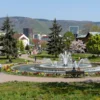 A panoramic view of Kislovodsk's central park in springtime. Focus on the lush greenery, blooming flowers, and the cascading water features. Include people strolling along the paths and enjoying the scenery. In the background, show the city's architecture and the surrounding mountains under a clear blue sky.
