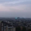 A wide shot of the city of Sumy at dusk. Buildings are visible, with a slightly hazy atmosphere suggesting smoke or dust. The sky is overcast with muted colors – grays, purples, and oranges. In the distance, faint glows or light flares can be seen, hinting at explosions. Focus on the urban landscape and the overall mood of alert and concern.