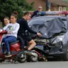 A photo-realistic image depicting a group of teenagers on a scooter colliding with a car in the village of Beloyarsk. The scene should show the aftermath of the accident, with the scooter and car damaged. Include emergency vehicles (police car and ambulance) arriving at the scene. Focus on the expressions of the teenagers and the seriousness of the situation.