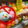 A close-up of colorful soft squishy toys shaped like cute animals and food on a store shelf, with a prominent red prohibition sign overlaid on them, a subtle toxic hazard symbol in the background, and a serious inspector's hand removing one from the display, highlighting the danger of carcinogenic substances in children's toys