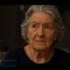 A somber, realistic portrait of an elderly woman with worried eyes, sitting in a dimly lit room. The room has a sense of old-fashioned domesticity, with antique furniture and muted colors. Focus on her expression of grief and fear.