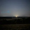 A wide shot of a nighttime landscape over the Kursk region, Russia. Silhouetted fields and forests stretch towards the horizon under a dark sky. A faint, distant glow suggests the aftermath of drone activity, perhaps with subtle streaks of light. Focus on the vastness of the landscape and the quiet stillness of the night.