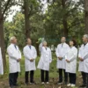 A natural outdoor setting with a group of scientists in lab coats standing in a forest clearing, releasing a swarm of small insects into the air for a sterilization experiment. The scene shows trees, grass, and a clear sky, with the insects depicted as tiny flying creatures. The atmosphere is scientific and calm.