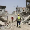 A wide shot of the aftermath of an earthquake in Venezuela. Buildings are damaged, rubble is scattered, and dust fills the air. Rescue workers are seen searching through the debris. The scene conveys a sense of devastation and urgency.