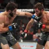 A dynamic and intense fight scene between Islam Makhachev and Garry Tonon. Makhachev should be in a dominant stance, showcasing his grappling skills, while Tonon is actively defending and attempting to counter. The background should be a blurred octagon with a roaring crowd. Focus on the athleticism and physicality of the fighters.