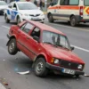 A damaged red Niva car overturned on a city street after a collision. Debris is scattered around the car. Emergency vehicles (police car and ambulance) are present in the background. The scene is captured during daylight hours.