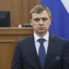 A serious-looking Russian man in a suit standing in front of a Russian flag, with a blurred background of a courtroom.