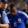 A soccer player from Côte d'Ivoire in a blue jersey being escorted by police officers in a stadium. The player looks distressed. Focus on the player's face and the surrounding police presence.