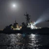 A covert nighttime rescue operation by American naval forces in the Strait of Hormuz: dark sea with faint moonlight, US Navy destroyer and a small support vessel illuminated by searchlights, Navy helicopters hovering, crew members lowering rescue ropes to a distressed merchant ship, subtle silhouettes of distant coastlines, atmospheric clouds of mist, realistic lighting and reflections on water