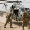 A military medical evacuation scene in a war zone. Several soldiers in uniform are assisting a wounded soldier being loaded onto a helicopter. Other soldiers are present, providing security. The environment is a dusty, outdoor area with military vehicles in the background. Focus on the urgency and teamwork involved in the evacuation.
