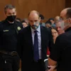 A courtroom scene in Cyprus. A man in a suit is being led away by security guards. The courtroom is filled with people watching. Focus on the man and the security guards.