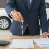 A person in a business suit holding a car key, standing in front of a car dealership with a surprised expression, surrounded by scattered documents and a calculator on the ground, symbolizing unexpected financial pressure