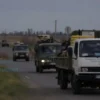 A solemn military transport convoy moving from Inzensk district toward a frontline zone, carrying humanitarian aid supplies and volunteers, set against a realistic Russian rural landscape with distant military vehicles and muted, serious atmosphere