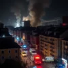 A wide shot of Odessa, Ukraine, at night. Buildings are damaged, with smoke rising from several locations. Emergency vehicles (ambulances, fire trucks, police cars) are visible in the streets, some with flashing lights. The overall atmosphere is one of chaos and destruction. Focus on the urban landscape and the immediate aftermath of the explosions.
