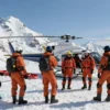 A group of rescuers on Mount Elbrus, wearing professional gear and equipment, standing near a helicopter on a snowy mountain peak. The rescuers are engaged in a search and rescue operation, with some carrying stretchers and others using radios. The background features the majestic snow-covered peaks of the Caucasus Mountains under a clear blue sky.