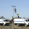 A repair crew restoring power lines in a rural area of Tula Oblast, Russia, with utility trucks, workers in high-visibility vests fixing damaged poles, and restored electricity flowing to nearby homes under a clear sky