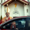 A church leader (priest or bishop) standing in front of the Holy Trinity Church, wearing traditional religious attire, with a surprised or shocked expression, as if he has just collided with a tourist driving an auto (car). The scene should be set in a realistic urban environment, with the church visible in the background, and the tourist car approaching or partially visible in the foreground.