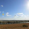 A realistic depiction of the Volgograd region in Russia on a summer afternoon, showing a bright sun, scattered clouds, a gentle breeze over the steppe and the Volga River, with subtle visual cues of a weather forecast such as faint temperature symbols integrated into the sky, without any written text or labels.
