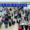 A bustling airport terminal with passengers walking, luggage carts, and airport staff assisting travelers. Include check-in counters, security checkpoints, and boarding gates in the background. Show a mix of people with suitcases, some rushing, others waiting. Add elements like digital flight displays, signage, and a sense of organized movement.