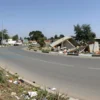 A damaged section of a recreational area near a roundabout in Senigal, with debris scattered around, broken pavement, and a partially collapsed structure. The scene should show a mix of urban decay and natural elements like overgrown grass or weeds. Include a clear sky and some distant trees or buildings in the background.