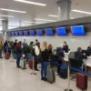 A busy airport terminal in Novosibirsk with long queues of passengers waiting at check-in counters, some holding luggage, while flight information screens display delays and cancellations. Airport staff in uniforms assist travelers, and digital boards show updated flight statuses.