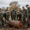 A realistic scene in a Russian town called Yartsevo, showing a group of cautious workers in protective gear examining a rusted, WWII-era explosive device partially buried in the ground near an old wooden fence, with autumn trees and a cloudy sky in the background, emphasizing the historical atmosphere and the seriousness of the find