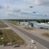An aerial view of Ulyanovsk Airport with closed runways, empty terminal buildings, and drones flying ominously in the sky above, surrounded by warning signs and emergency vehicles on the ground, under a cloudy sky