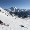 A panoramic view of Mount Elbrus in the Caucasus Mountains during late spring or early summer, showing remnants of ski equipment on snow-covered slopes, possibly a few skiers or snowboarders in the distance, clear blue sky, alpine landscape with rocky ridges and distant valleys, natural lighting, no text or numbers on the image